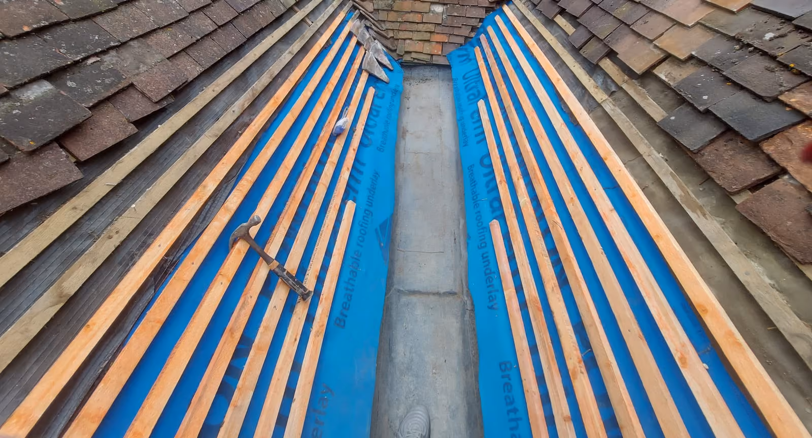 A roof valley stripped back, with new battens laid over blue breathable underlay on both slopes